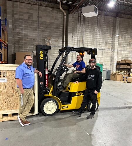Three workers posing by a forklift in a warehouse smiling at the camera.